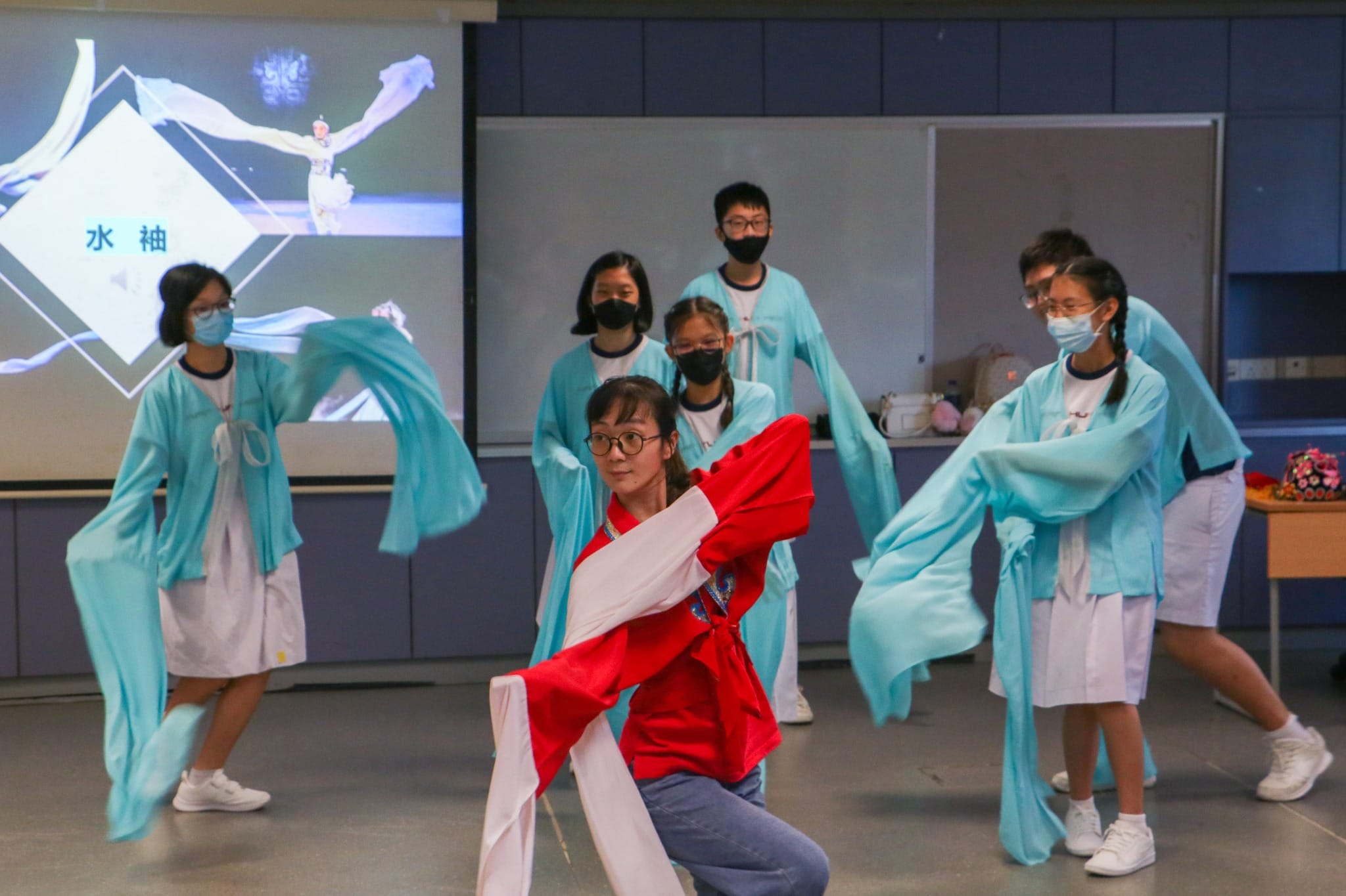 Students in teal robes with flowing sleeves perform a traditional Chinese water sleeve dance in a classroom.